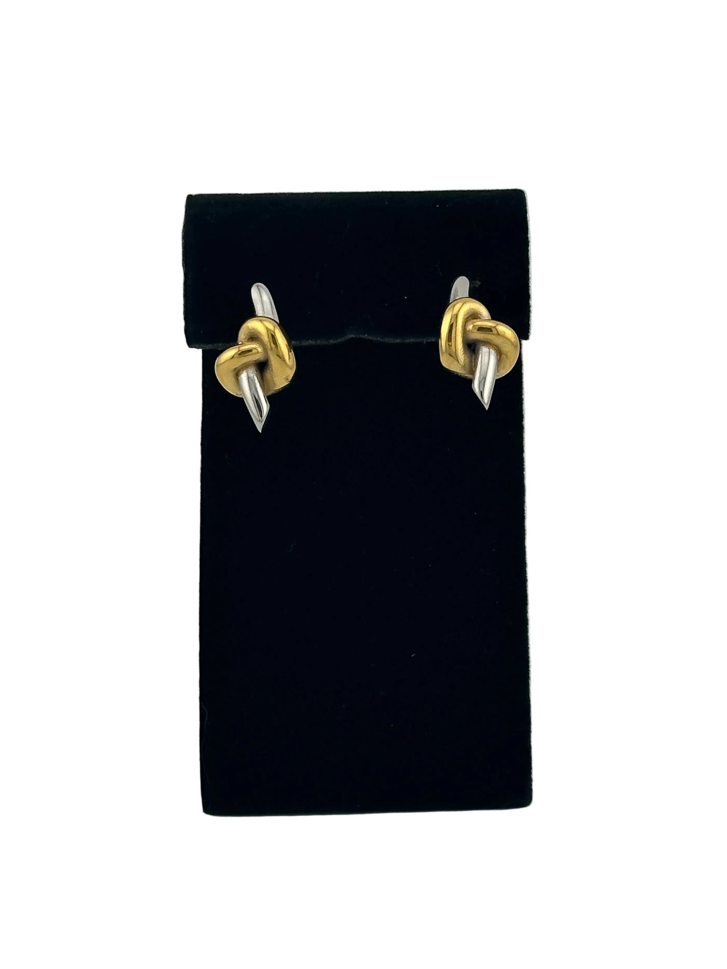 A pair of small gold and silver knot stud earrings displayed on a black rectangular jewelry stand against a light gray background.