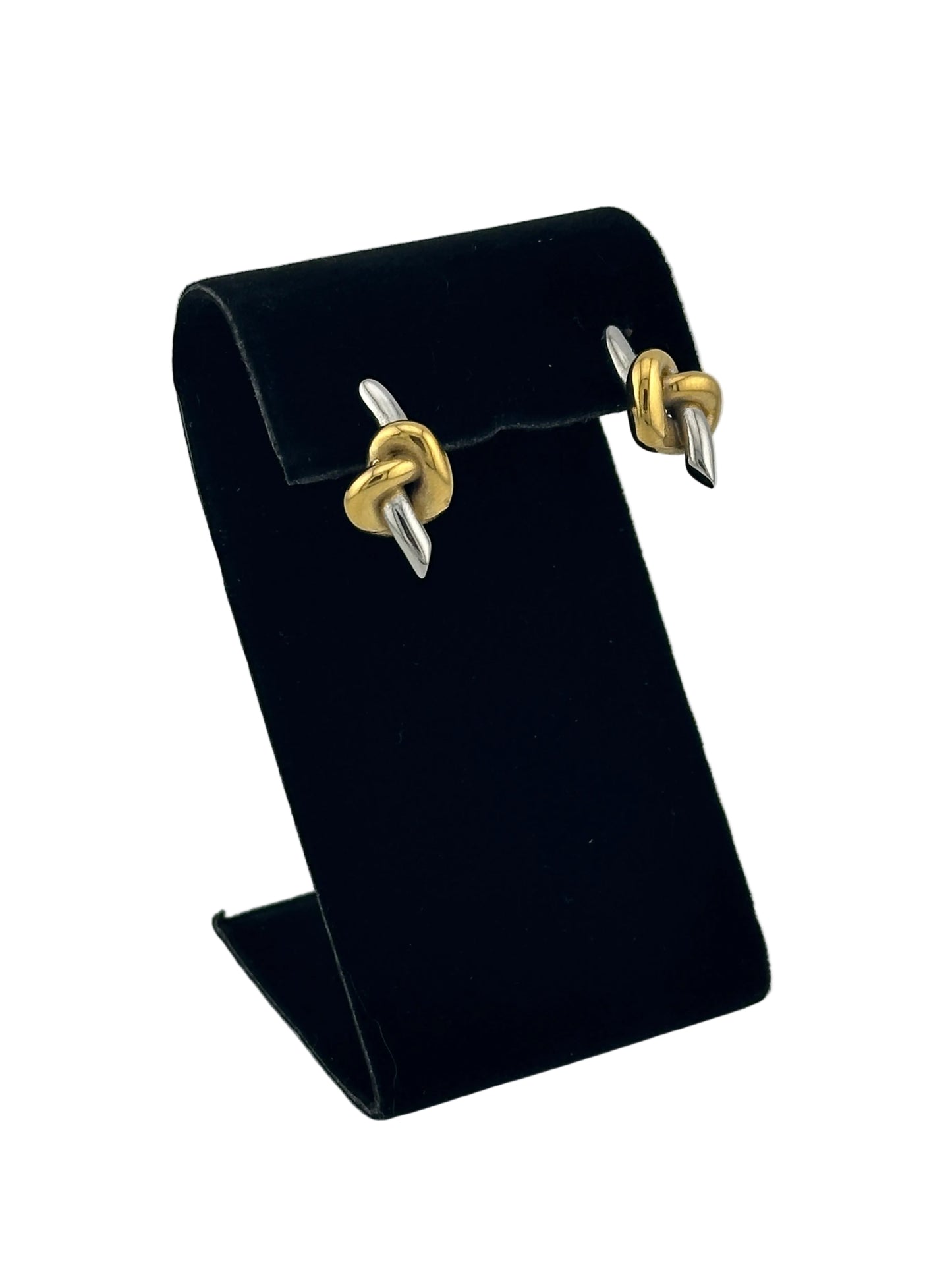 A black velvet jewelry display stand holding a pair of gold and silver knot stud earrings, photographed on a light gray background.